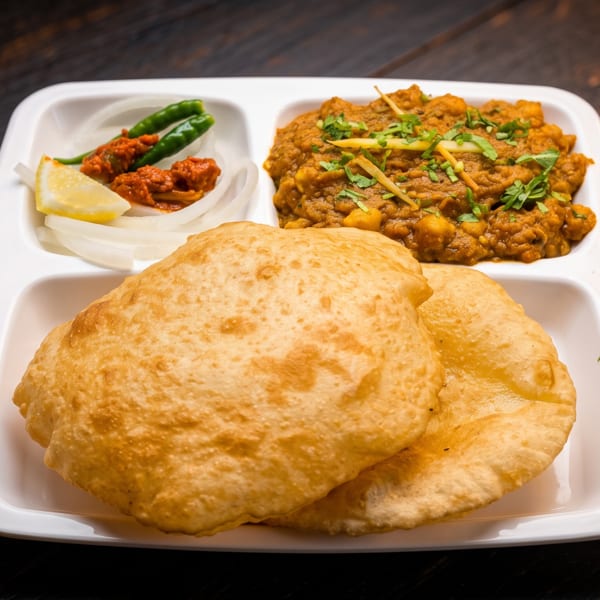 Indian meal with puffed fried breads, chickpea curry, chutney, green chilies, lemon, and sliced onions on a white plate.