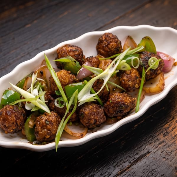 Crispy meatballs with sautéed peppers and onions, garnished with sliced green onions on a white platter.