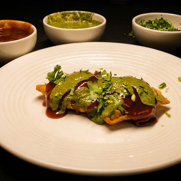 Samosas with red and green chutneys on a white plate, accompanied by three sauce bowls.