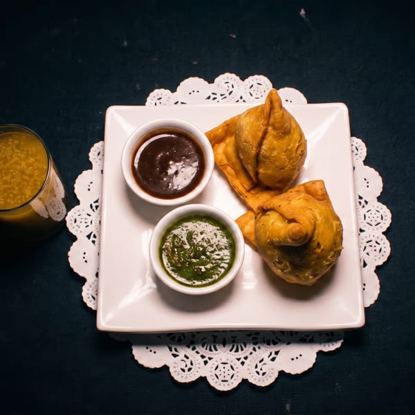 Two golden samosas with green and brown chutneys on a white plate, accompanied by a glass of yellow beverage.