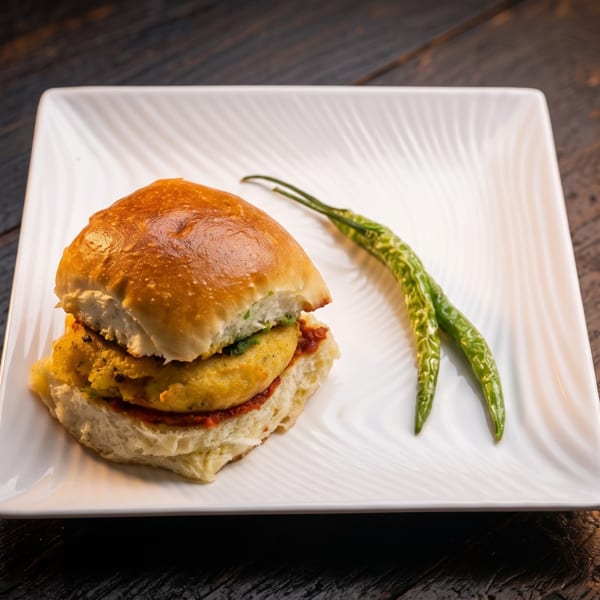 A golden-brown stuffed sandwich with a crispy filling sits on a white plate beside two green chilies.