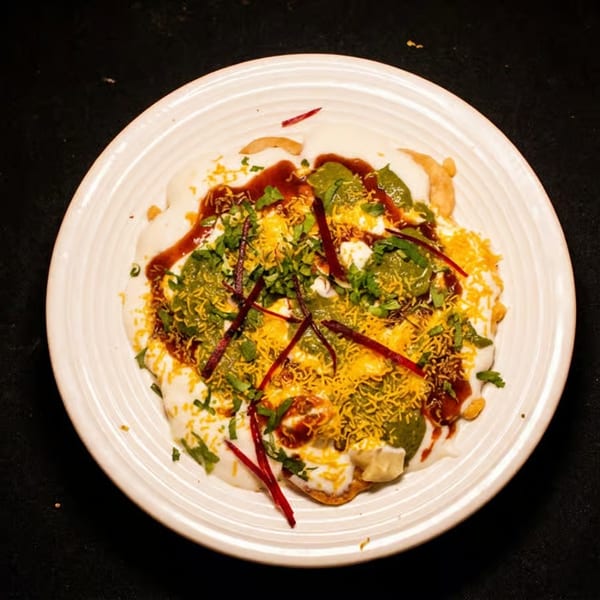 Indian chaat platter topped with yogurt, chutneys, crispy sev, chopped herbs, and beet strips on a white plate.